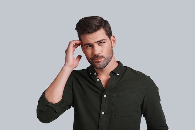 Feeling uncertain. Thoughtful young man touching his head with hand and looking away while standing against grey background