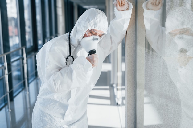 Feels bad and sick. Female doctor scientist in lab coat, defensive eyewear and mask standing indoors