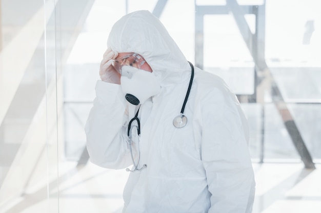 Feels bad and sick. female doctor scientist in lab coat, defensive eyewear and mask standing indoors