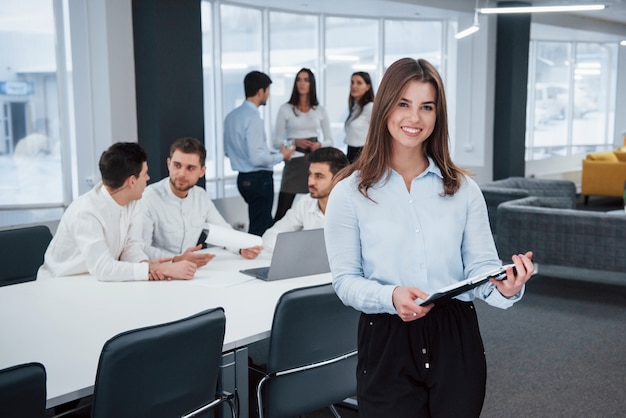 Foto si sente bene perché ama il suo lavoro. il ritratto della ragazza sta nell'ufficio con gli impiegati a fondo