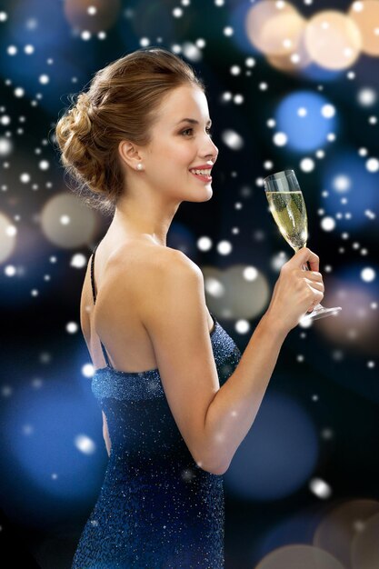 feest, drankjes, vakantie, kerstmis en mensenconcept - glimlachende vrouw in avondjurk met glas mousserende wijn over nachtverlichting en sneeuwachtergrond