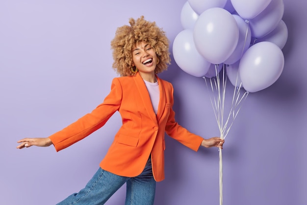 Feest is op Vrolijke vrouw gekleed in modieuze oranje jas en spijkerbroek houdt stelletje opgeblazen ballonnen heeft vrolijke expressie feestelijke stemming geïsoleerd over paarse achtergrond Speciale gelegenheid