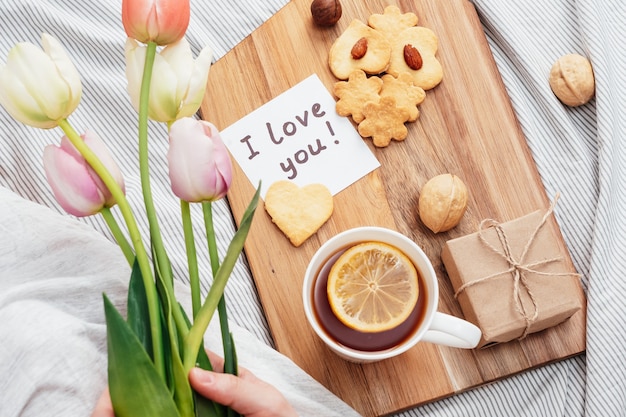 Feestelijk ontbijt op bed voor Valentijnsdag. Thee en koekjes met je eigen handen in de vorm van harten. Een briefje op papier, een cadeautje en bloemen voor je geliefde meisje.