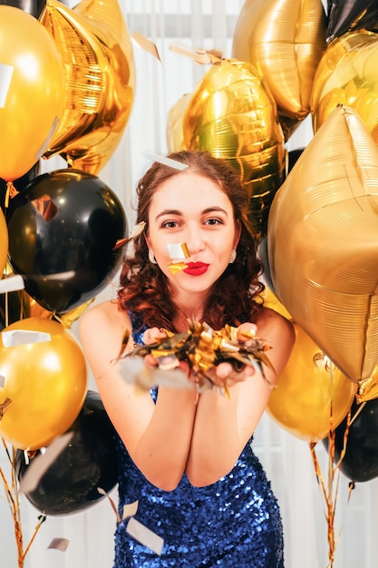 Feestelijke gelegenheid Close-up portret van brunette meisje in sprankelende jurk die confetti blaast tussen gouden en zwarte ballonnen