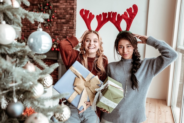 Feestelijke manier. Aantrekkelijke langharige meisjes die feestelijke hertenoren en versierde geschenken tonen terwijl ze in de buurt van de kerstboom staan