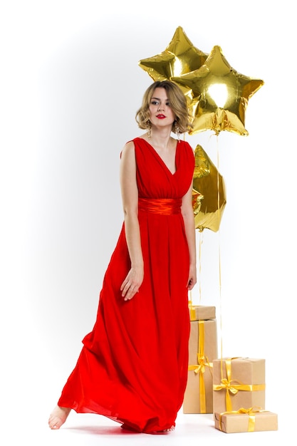 Feestfoto van elegantie sexy dame in rode jurk met rode lippen en blond mooi krullend haar, glimlachend. Achtergrond van gouden luchtballonnen en geschenken. Concept van de verkoop