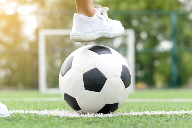 Feet of a boy wearing white sneakers stepping on a soccer ball in the middle of the football field.