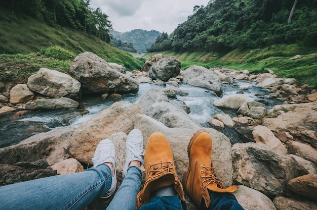 I piedi delle coppie che si rilassano mentre sedendosi guardano la vista del fiume della montagna