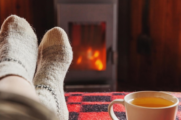 Feet legs in winter clothes wool socks and cup tea at fireplace background woman sitting at home on
