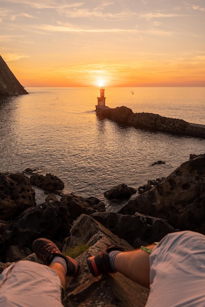 San juan gipuzkoa basque country の町の faro de la bahia de pasaia でオレンジ色の夕日を見て座っている男の足