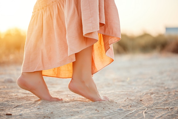 Feet of a woman in a pink dress walking on the sand during sunset