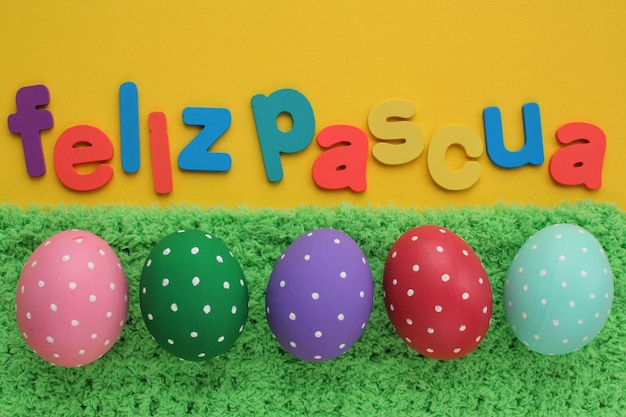 펠리즈 파스쿠아(Feliz Pascua), 노란색 배경에 스페인어로 된 행복한 부활절 편지와 녹색 잔디에 색 계란. 호