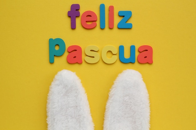 Feliz Pascua, Happy Easter on spanish colored lettering and white bunny fur ears on yellow backgroun