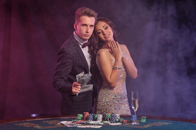 Fellow is holding cash playing poker at casino at table with stacks of chips and cards on it He celebrating win with gorgeous lady Black smoke background colorful backlights Gambling Closeup