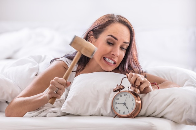 Female about to hit alarm clock with a hammer while lying in white sheets in her bed