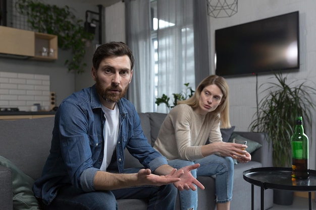 Female alcoholism husband in despair woman wife drinking hard alcohol couple sitting on sofa in living room