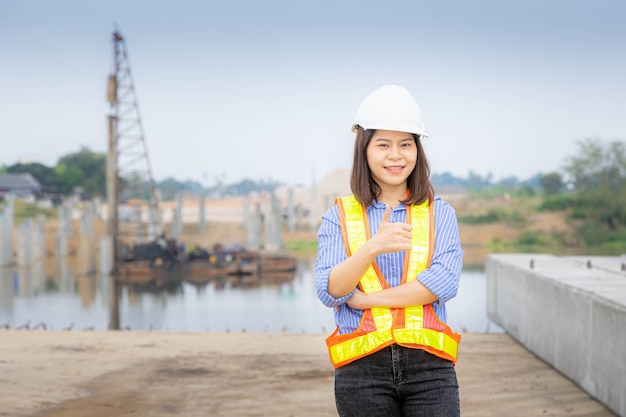 A female architect leader standing at the bridge construction,  Wearing a safety helmet and holding the blueprint of the project