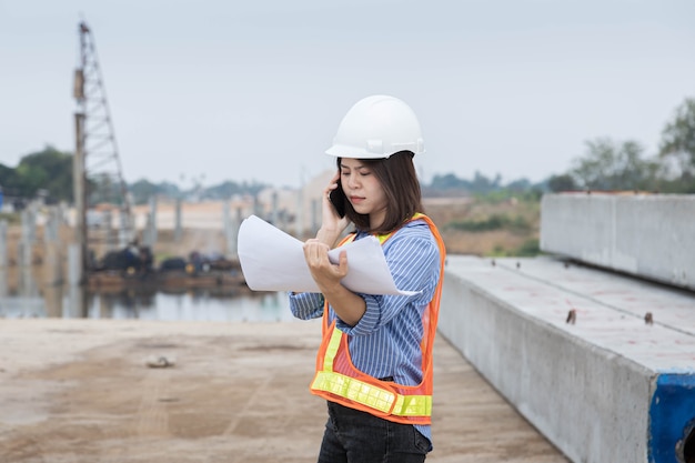 A female architect leader Wearing a safety helmet are using smartphone to check blueprint of bridge construction.