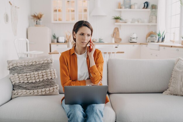 Female arguing by phone dissatisfied with bad customer support\
sitting with laptop on sofa at home