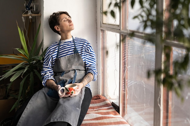 Female artisan holding cup of tea sitting on windowsill taking break during workday in workshop