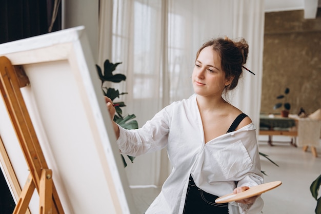 Female artist in casual clothes sitting on chair in front of easel and painting with brush and color palette