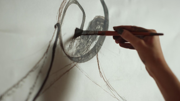 Female artist hand painting figure in black with paintbrush in art workshop Unknown young woman working on masterpiece indoors Closeup painter hand holding brush on white background