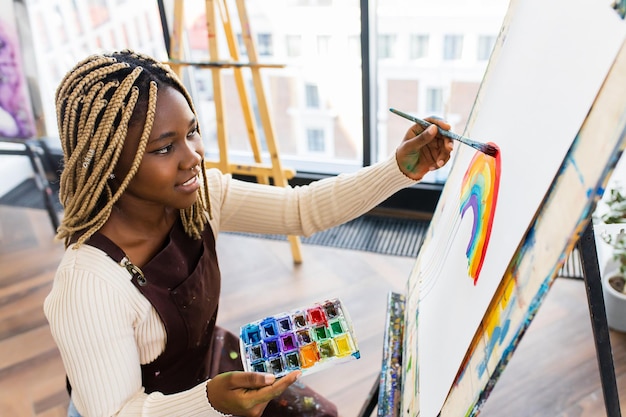 Female artist with brush and paint palette drawing rainbow in workplace art class