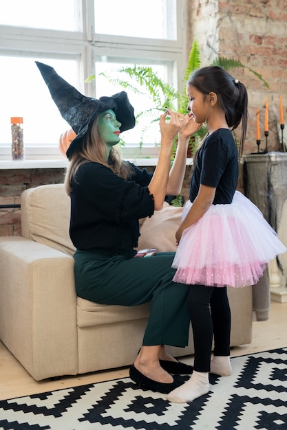 Художница с зеленым лицом в шляпе ведьмы делает макияж на Хэллоуин азиатской девушке, готовя ее к праздничной вечеринке