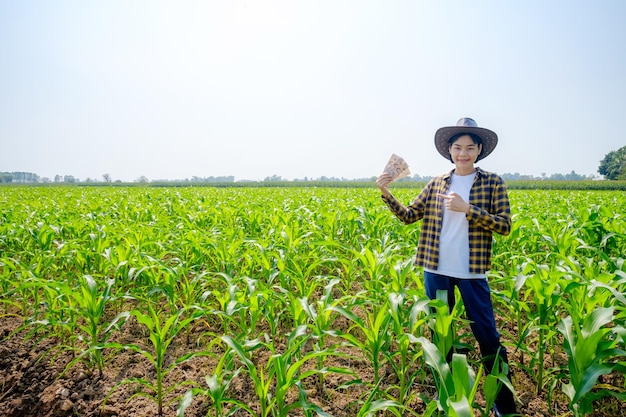 A female Asian farmer in a striped shirt wearing a hat posing standing with corn income bills