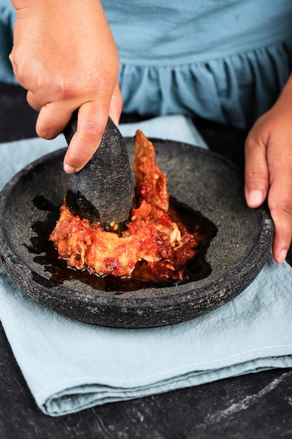 인도네시아의 인기 있는 길거리 음식인 아얌 게프렉(Ayam Geprek)을 만들거나 요리하는 과정에서 인도네시아 모르타르와 페슬을 곁들인 매운 칠리 페이스트를 곁들인 여성 아시아 손으로 으깬 프라이드 치킨.