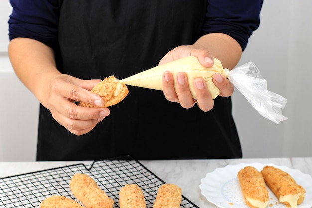 Femmina asiatica a mano ripieno di pasticceria francese craquelin eclair profiterolle con crema utilizzando la sacca da pasticceria. dolci casalinghi preparazione cottura di pasta choux / bignè alla crema.