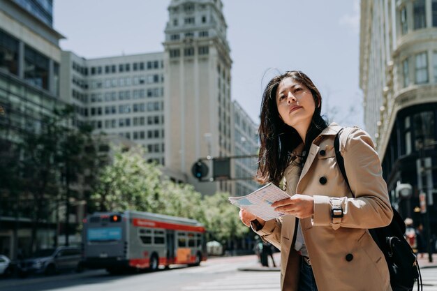 Female Asian Tourist Exploring City While Holding Paper Map On Sunny Day. Young Girl Traveler With Backpack Self Trip In San Francisco Usa. Car Public Transport Bus Light Railway On Road.