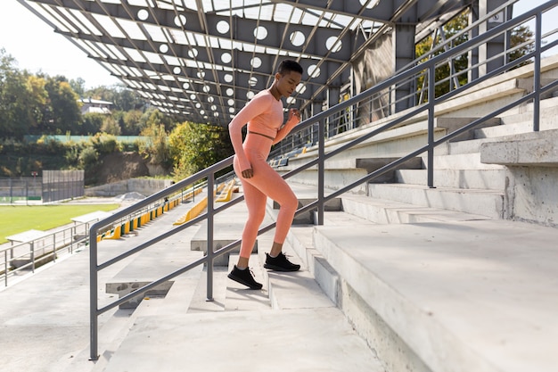 Female athlete doing fitness in a pink tracksuit near a sports stadium, African American running up the stairs in the morning