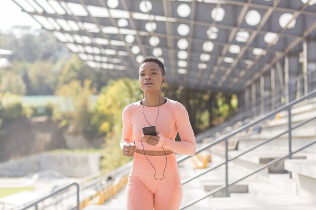 Female athlete doing fitness in a pink tracksuit near a sports stadium, African American running up the stairs in the morning