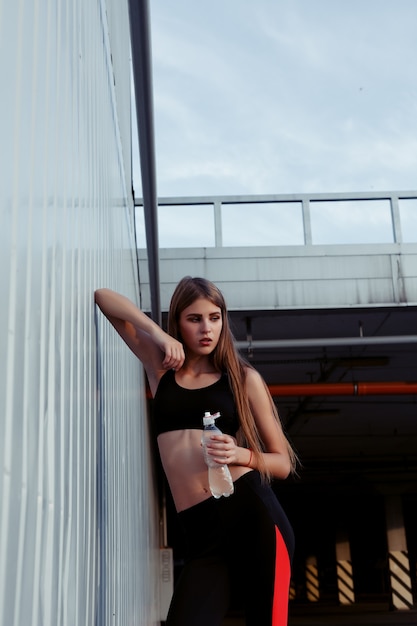 Female athlete drinking water while standing by a grey wall. Slim woman in sportswear taking a break after workout.