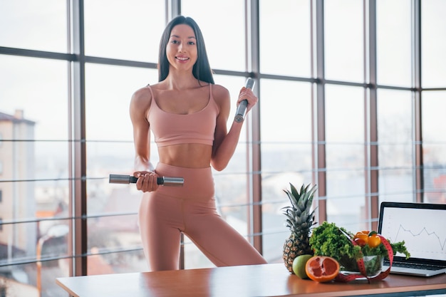 Female athlete in sportive wear holds dumbbells and standing near table with laptop and healthy food.