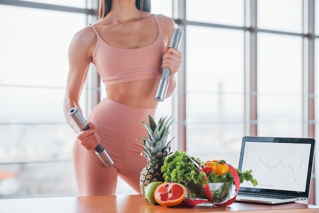 Female athlete in sportive wear holds dumbbells and standing near table with laptop and healthy food.