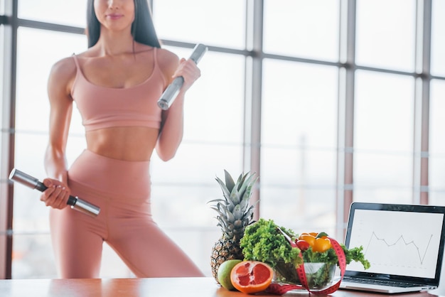 Female athlete in sportive wear holds dumbbells and standing near table with laptop and healthy food.