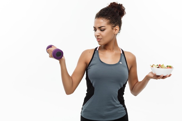 Female athlete with food and dumbbell standing against white background