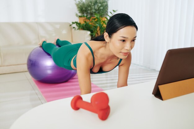 Female athlete working out at home, doing plank exercise with legs on fitness ball