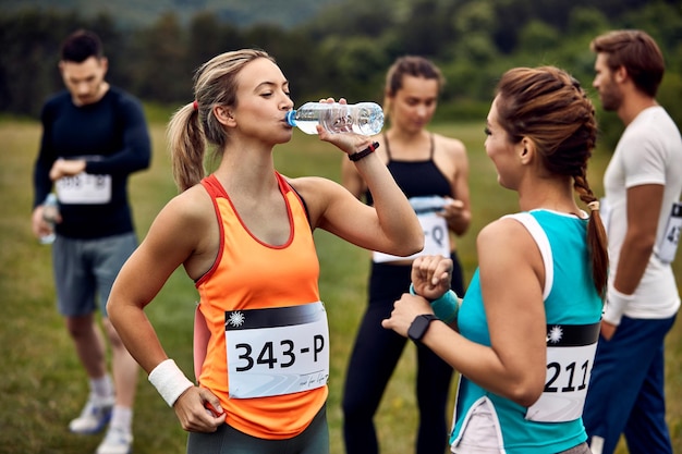 자연 속에서 마라톤 경주에서 휴식을 취하는 동안 의사 소통하는 여성 운동 선수는 병에서 물을 마시는 여성에 초점을 맞추고 있습니다