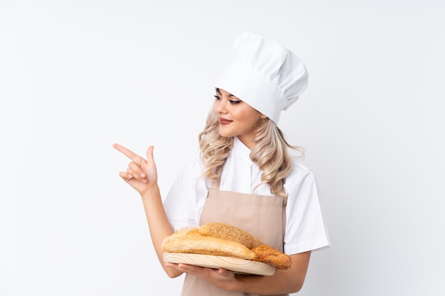 Female baker holding a table with several breads over isolated white background pointing finger to the side