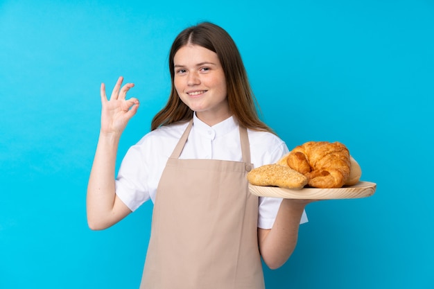 Female baker holding a table with several breads showing ok sign with fingers