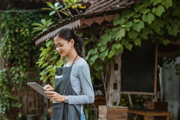 Female barista in apron looking at the digital tablet while standing outside the coffee shop