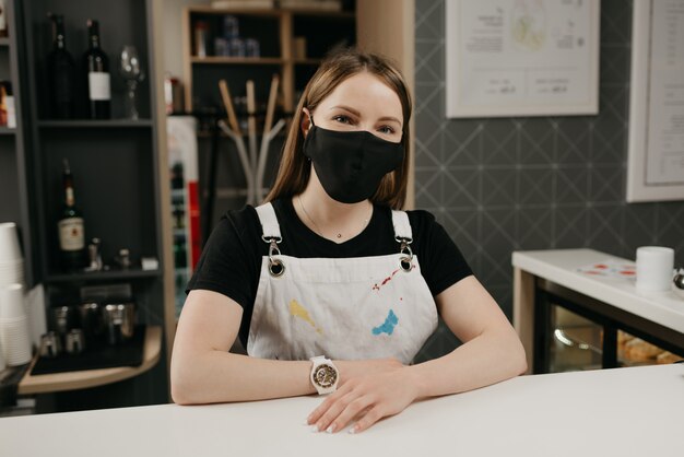 A female barista in a medical black face mask smiles and waiting for clients in the coffee shop