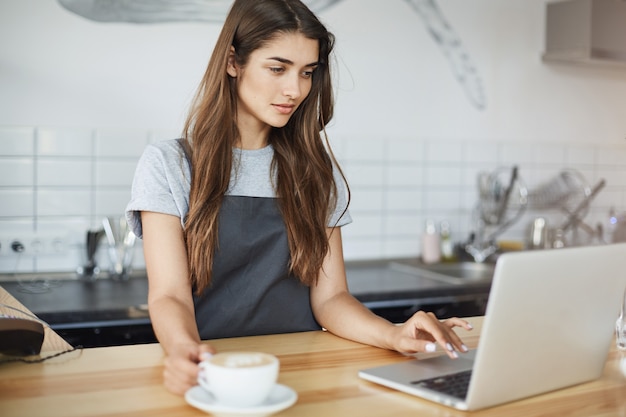 Female barista running her coffee business having a small break surfing web and social media on a laptop.