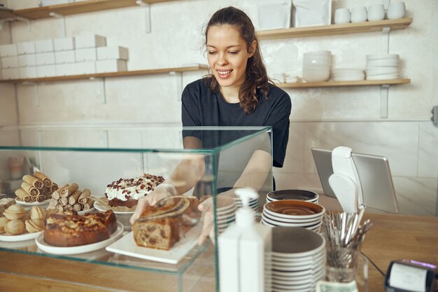 Female barista at showcase with desserts in cafe. Young worker of sweet shop and cafe.