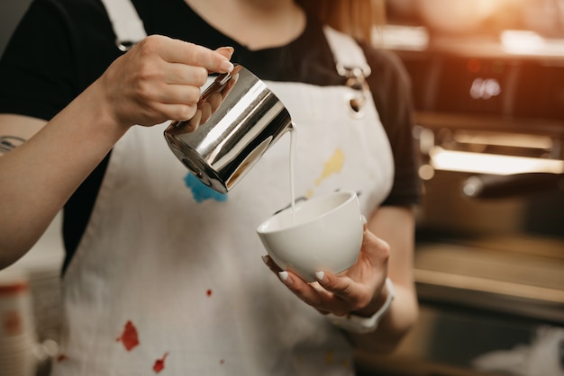 A female barista with a metal mug of milk crafts a latte art on a coffee in a cafe. A barista pouring milk with a narrow stream to avoid breaking the espresso's crema.