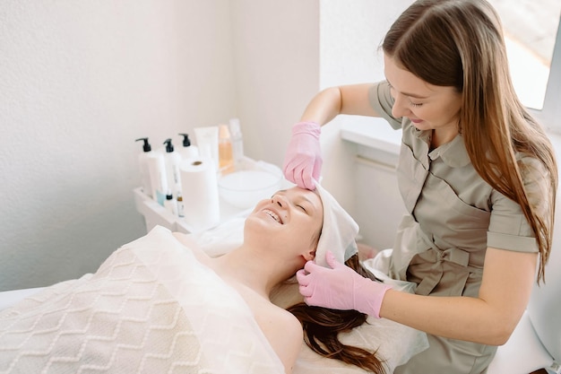 Женщина-косметолог прячет волосы от лица молодой девушки, готовящей ее к косметическим процедурам