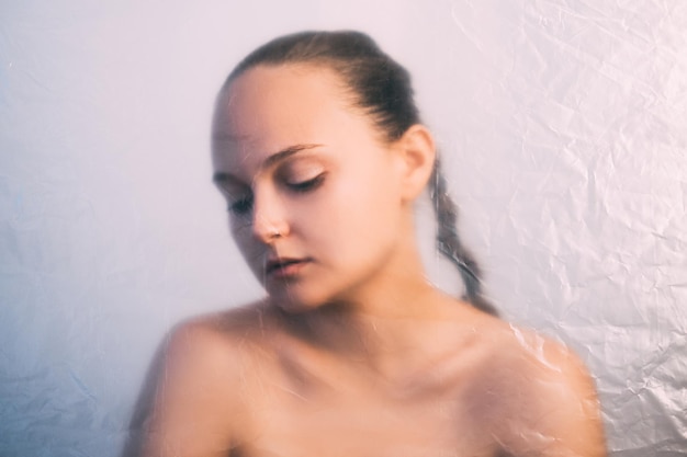 Photo female beauty. defocused woman portrait. skincare detox. wrinkled texture lady silhouette with nude makeup bare shoulders behind transparent creased film isolated on light background out of focus.